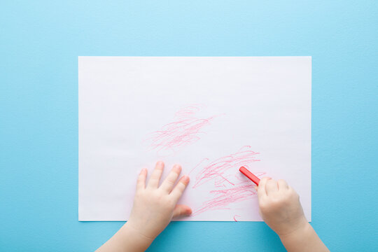 Baby hand holding red wax crayons and drawing first scratches lines on white paper on light blue table background. Pastel color. Closeup. Toddler development. Point of view shot. Top down view.