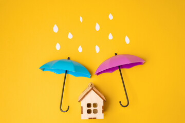 A toy umbrella over a wooden house on a yellow background. A symbol of care and reliability