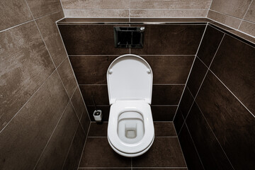 Ceramic toilet bowl with toilet seat, chrome wall button and toilet brush in restroom. Paving and tiling have texture of concrete trowel in shades of brown color. The toilet seat is open.
