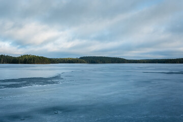 Frozen lake in wilderness with forest on horizon and sky with floating clouds. Ice has soft melting structure, trees in background are slightly illuminated by sun and blue sky shines through clouds.