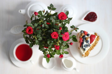 Red roses flowers on white table with tea crockery set and cake with berries flat lay