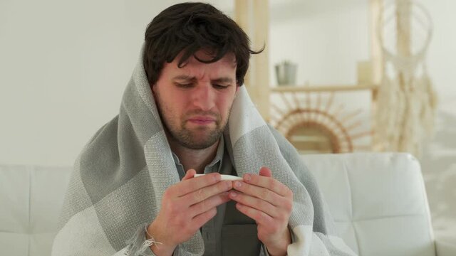 Man with a cold is sitting on the couch with a thermometer in his mouth. A person at home is sick with the flu, measures his temperature