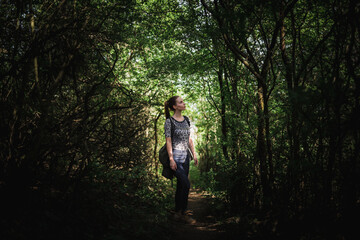 Young woman standing in middle of dense vegetation forming narrow alley. Soft light shines through vault of the branches and leaves. Woman is casually dressed with hydrating backpack on her back.