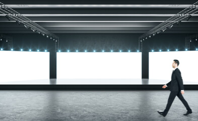 Businessman walks in empty exhibition hall with three glowing blank white screens, light on top, black podium and concrete floor