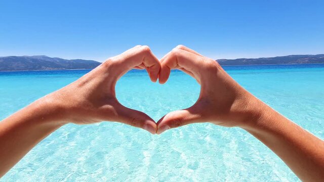 Girl loving tropical paradise. Young woman on top of idyllic beach makes a heart shape with hands showing love to nature and environment- slow motion 