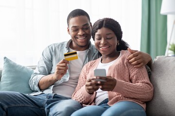 Happy black wife and husband shopping from home, using smartphone