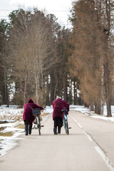 Fototapeta na wymiar Two senior women in red coats with old rustic bikes walk down the road in winter. Country fashion