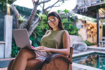 Smiling tanned female freelancer using laptop at poolside in tropical resort