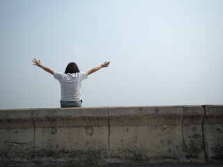An Asian woman sat at a river on the dam and happily raised her hand.