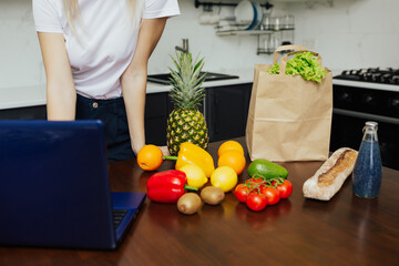 Online purchase. Cropped shot of  woman shopping with laptop in kitchen with paper bag full of vegetables nearby.