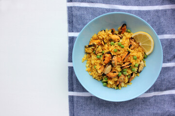 Traditional Spanish seafood dish paella with lemon slice on table. Mussel risotto with green peas in blue bowl on striped cloth napkin background. Top view, copy space. Healthy eating concept
