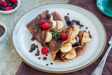 Thin chocolate pancakes stuffed with bananas, raspberries and blueberries, poured with chocolate sauce, on a gray ceramic plate on a light concrete background. Pancake recipes.