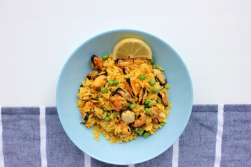 Traditional Spanish seafood dish paella with lemon slice on table. Mussel risotto with green peas in blue bowl on striped cloth napkin background. Top view, copy space. Healthy eating concept
