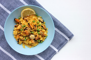 Traditional Spanish seafood dish paella with lemon slice on table. Mussel risotto with green peas in blue bowl on striped cloth napkin background. Top view, copy space. Healthy eating concept
