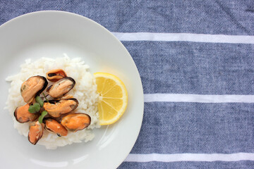 Mussel risotto on white plate on table. Top view of white rice with seafood and lemon slice on striped cloth napkin background. Copy space. Healthy eating concept
