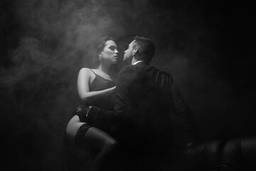 Monochrome shot of sensual woman standing near boyfriend in suit on black background with smoke