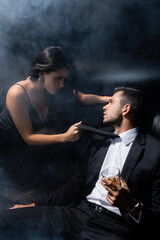 Brunette woman pulling tie of boyfriend with whiskey on black background with smoke