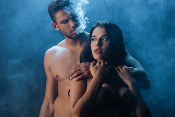 Sexy man smoking and hugging girlfriend in bra on black background with smoke