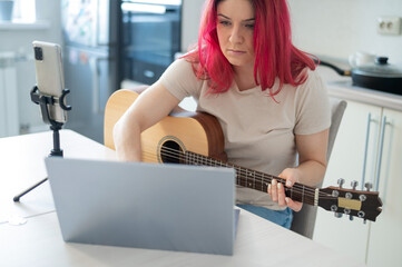 A woman sits in the kitchen during a remote acoustic guitar lesson. A girl learns to play the guitar and watches educational videos on a laptop