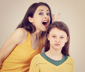 Happy laughing crazy mother showing the horns above her daughter head with angry unhappy face. Funny Family playing together on blue light background. Closeup