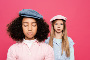 fashionable girl looking at camera near african american friend with closed eyes isolated on pink