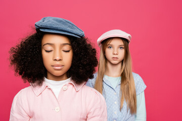 interracial girls in stylish clothes and caps posing isolated on pink