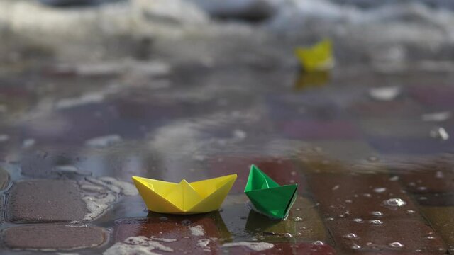 Early spring landscape. Melting winter snow turning into much water on sidewalks. 4k stock video footage of three cute small paper yellow and green boats floating on surface of spring water puddle.