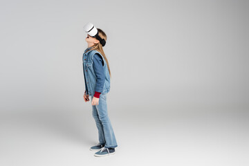 full length view of amazed girl in stylish denim clothes and vr headset standing on grey