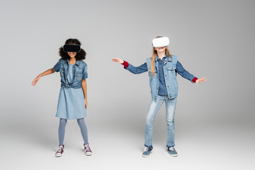 full length view of cheerful interracial friends in trendy denim clothes gaming in vr headsets on grey