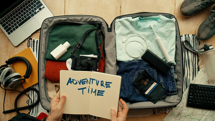 Man with paper notebook that says Adventure Time packs suitcase for international abroad travel. Neatly organized personal belongings. Top view social media concept. Personal items in bag