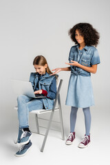 smiling, trendy african american girl pointing at friend sitting on chair and using laptop on grey
