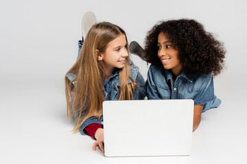 happy interracial girls in trendy denim clothes smiling at each other while lying on grey near...