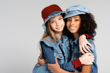 joyful interracial girls in stylish denim clothes embracing isolated on grey