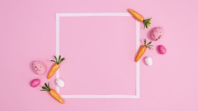 Easter eggs and carrots move around copy space frame on pastel pink background. Stop motion flat lay