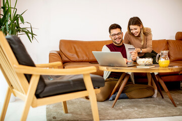 Young woman and young man using laptop for onlin payment while sitting by sofa at home