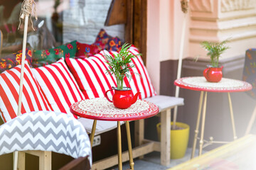 a red round table with a tablecloth and a flowerpot in the form of a teapot with a green plant by a chair with pillows, an outdoor cafe with empty seats, nobody.