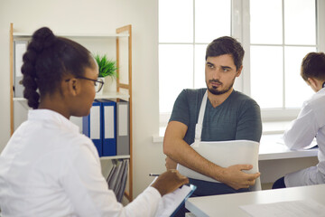 Fototapeta premium Caucasian man with hand in bandage sling pointing to injured elbow complains of arm pain at doctor appointment. African american woman specialist listening carefully to patient with hand trauma