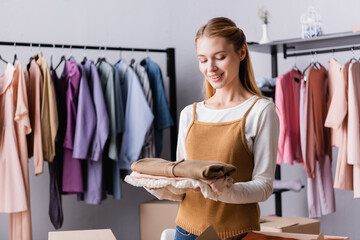 Happy showroom proprietor holding clothes near hangers on blurred background