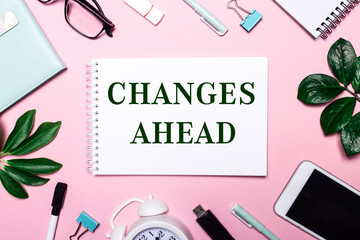 CHANGES AHEAD is written in a white notebook on a pink background surrounded by business accessories and green leaves.