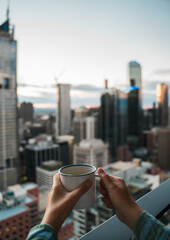 Junge Frau trinkt Kaffe auf einem Balkon und genießt die Aussicht über eine Großstadt...