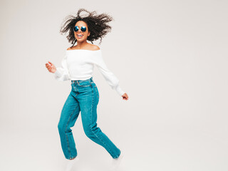 Full length of beautiful black woman with afro curls hairstyle.Smiling model in sweater and trendy jeans clothes. Sexy carefree female jumping on white background in studio. Dynamic movement