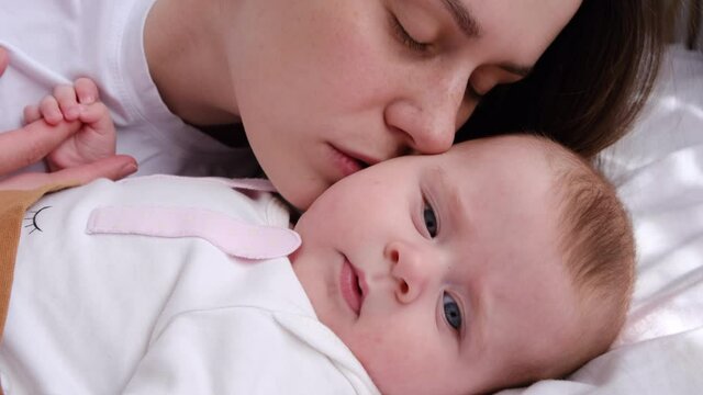 Loving beautiful young mum kissing on cheek cute adorable funny little kid daughter lying on comfortable white bed at home, happy family mother and small baby girl enjoying bonding having fun