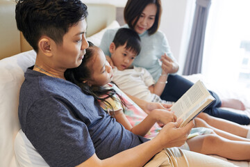 Vietnamese man reading interesting book for his children and wife whe they all are lying on bed
