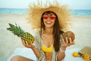 Joyful woman in red eyeglasses wearing straw hat smiling to camera on the beach, holding tropical fruit, girl looking happy on summer vacation.