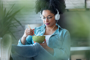 Beautiful woman eating noodles with chopsticks while listening music with headphone of work sitting...