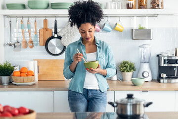 Beautiful afro woman eating noodles with chopsticks while standing in the kitchen at home.