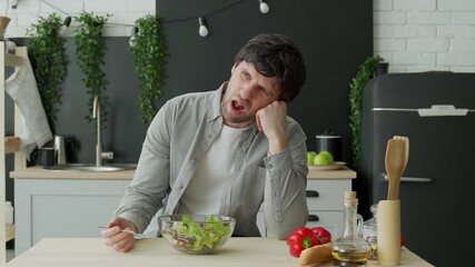 Unhappy man eating vegetable salad at table in kitchen. Displeased young man eating green leaf lettuce 