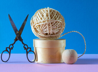 ball of string on a wooden pedestal with a pair of ornate scissors on a blue and pink background , concept of hobbys  and pastimes .