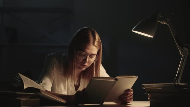 Late study. Night insomnia. Nerd lifestyle. Exhausted tired intelligent smart female student in eyeglasses fell asleep reading book late at dark library workplace with desk lamp light.