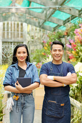 Naklejka premium Happy young Vietnamese man and woman with digital tablet standing at greenhouse they are working at and smiling at camera